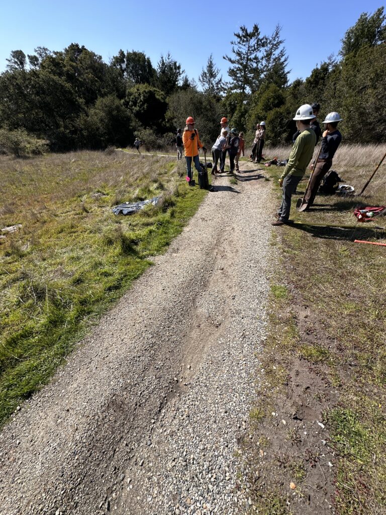 People standing on a trail about to repair. Edges of trail have green grass trail is rocks and brown